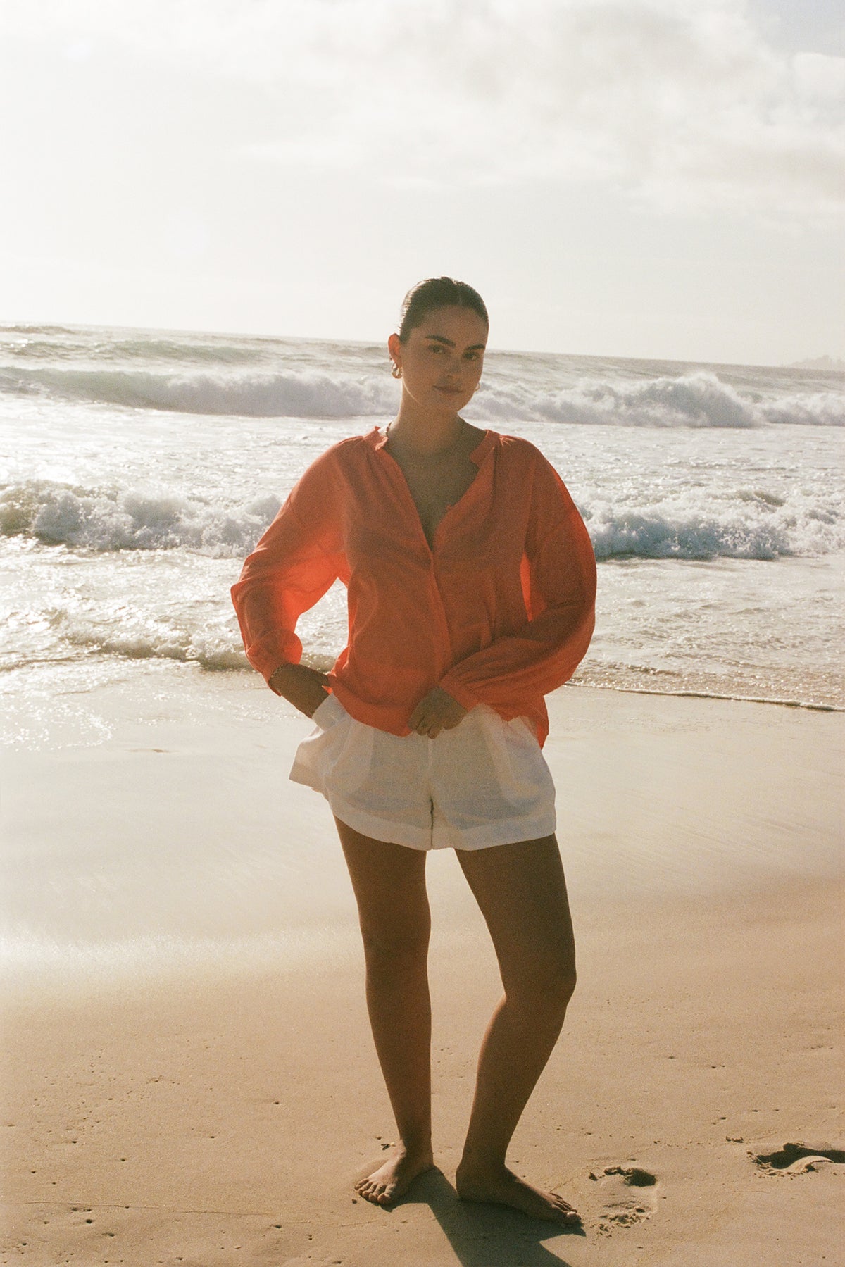 LILLY PILLY woman in a spritz Ember cotton voile top sitting and ivory white   Gaia shorts standing on a beach with waves in the background
