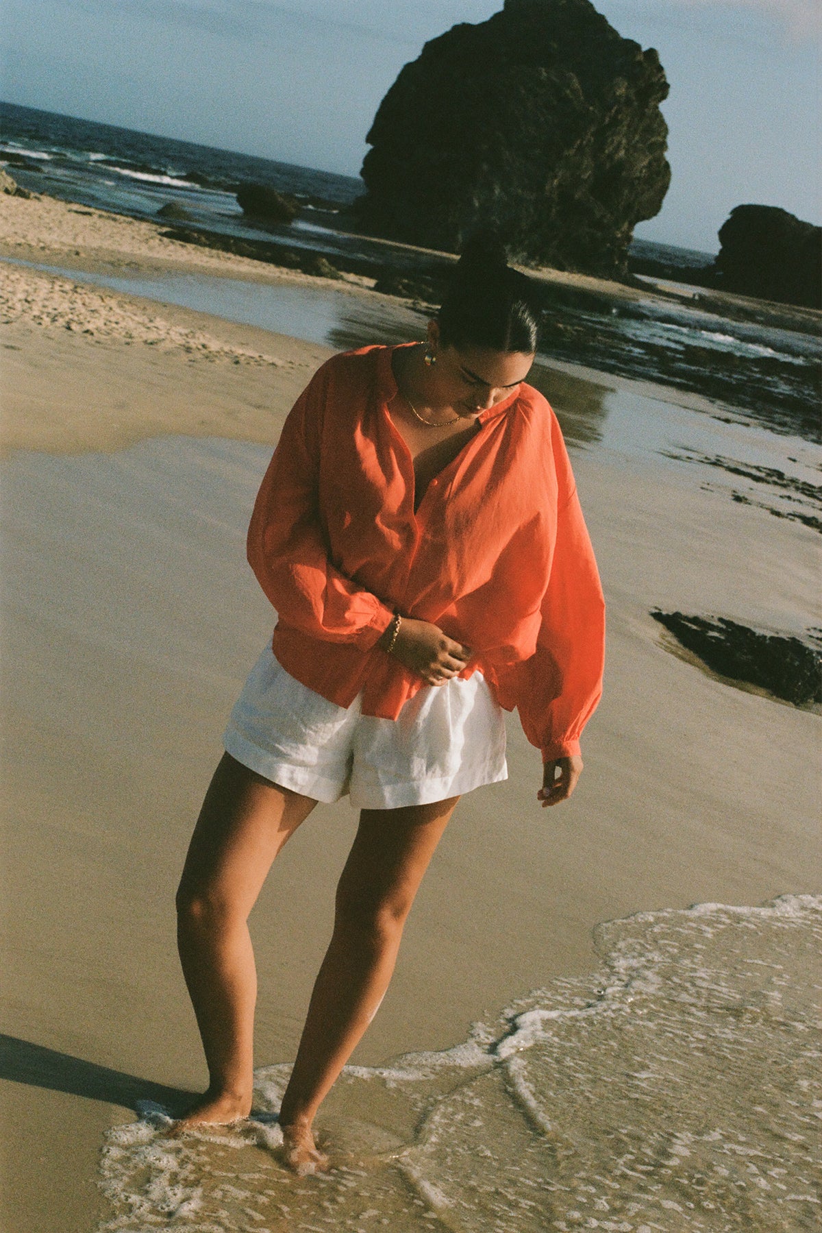 LILLY PILLY woman in a spritz Ember cotton voile top and white shorts standing on a beach with rocks and water in the background.