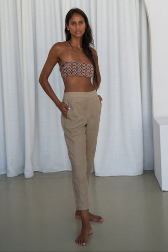Emer Linen Pants in Taupe - full length front view, woman wearing Taupe tapered linen pants with printed bandeau top, white curtain backdrop - Lilly Pilly Collection