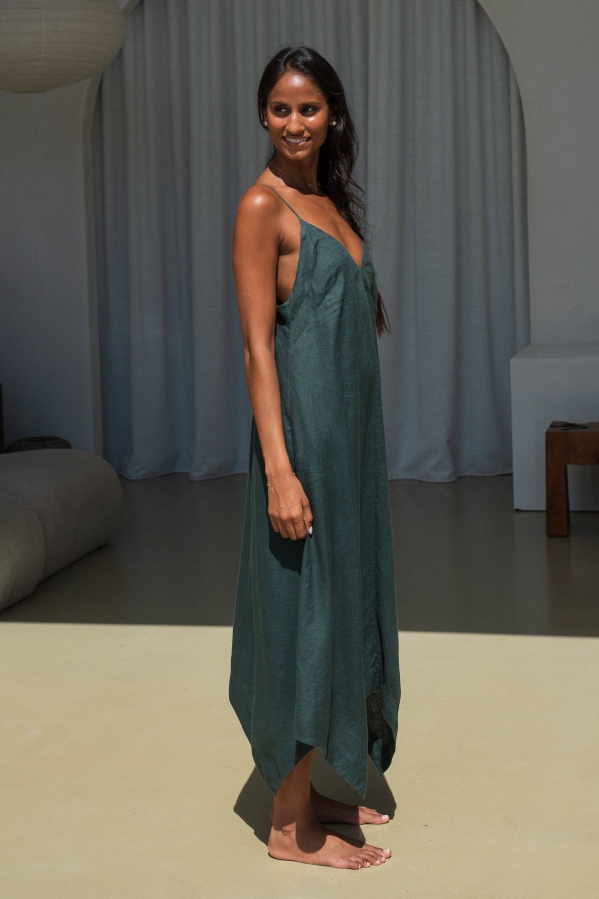 Side view of LILLY PILLY woman wearing a forest green Inez linen dress in a minimalistic room  with gray curtains.