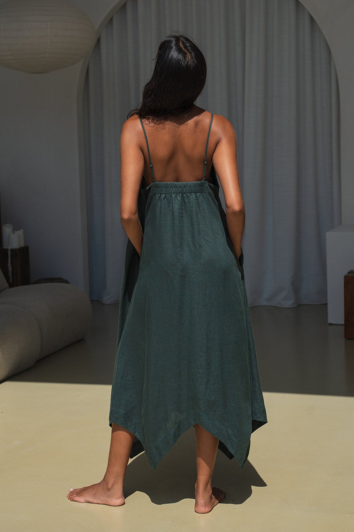 Back view of LILLY PILLY woman wearing a forest green Inez linen dress  standing in a room with neutral decor.