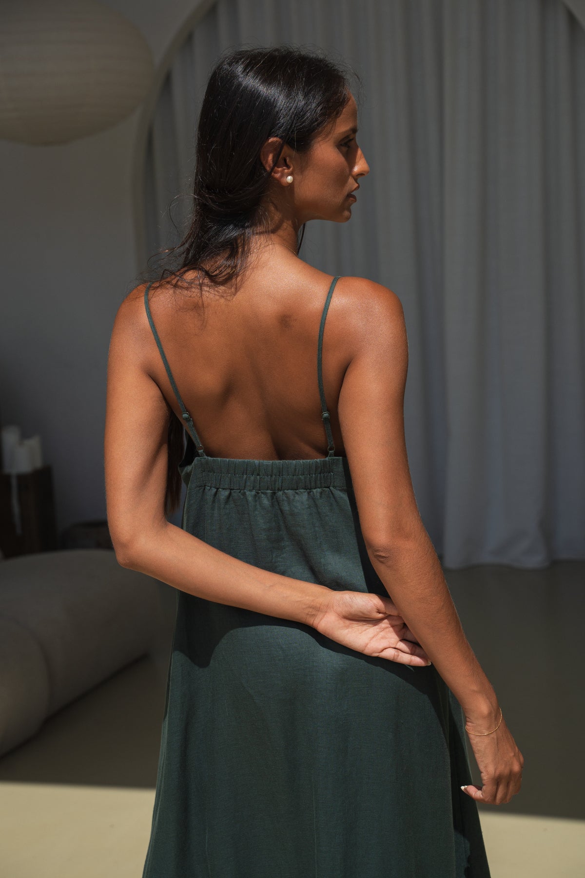 Close up back view  of LILLY PILLY woman wearing a forest green Inez linen dress  with thin straps in an indoor setting