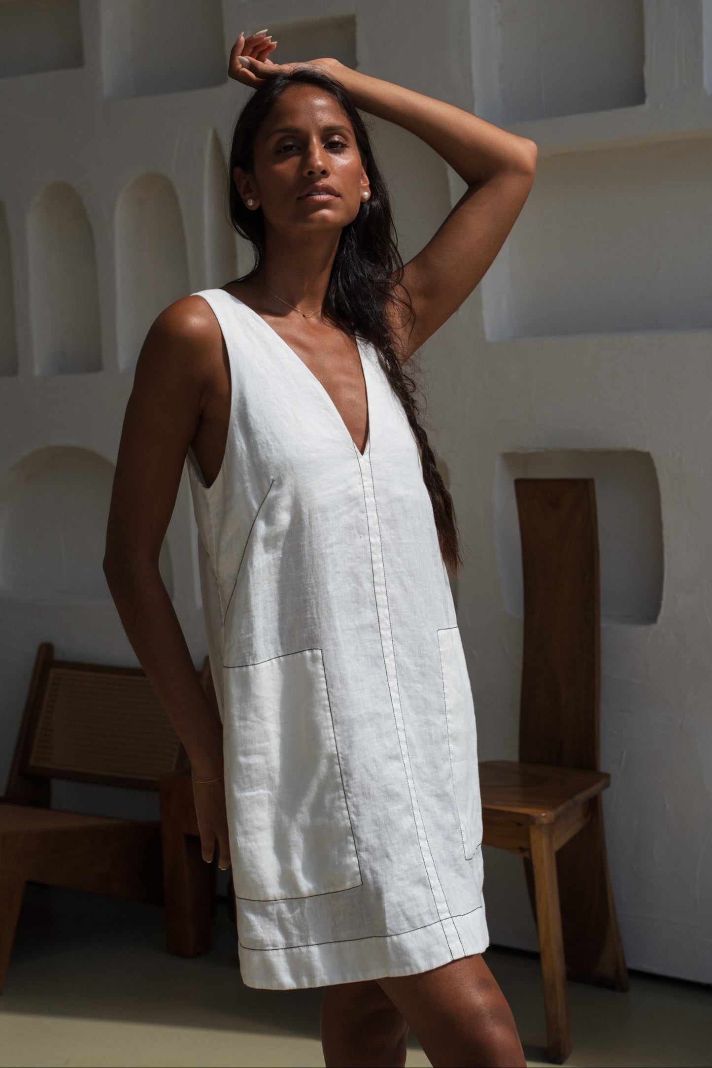 LILLY PILLY woman in an ivory Isla linen dress standing in a minimalistic setting with wooden chairs.