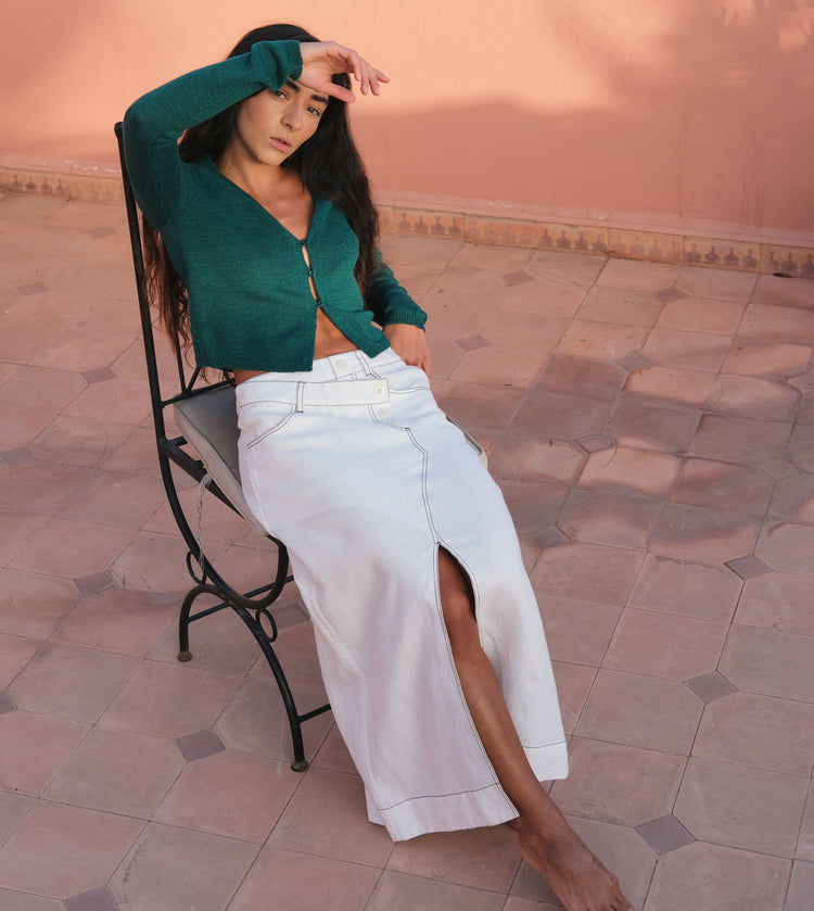 LILLY PILLY woman sitting on a chair wearing a oine green Indie cardigan  and ivory Jax linen skirt with a blurred background