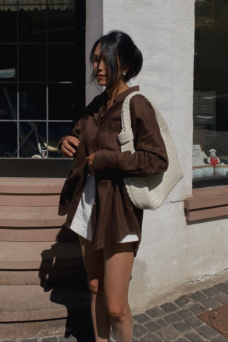 Kirra Linen Shirt in Chocolate - side profile, holding woven bag, worn open over shorts, street setting - Lilly Pilly Collection
