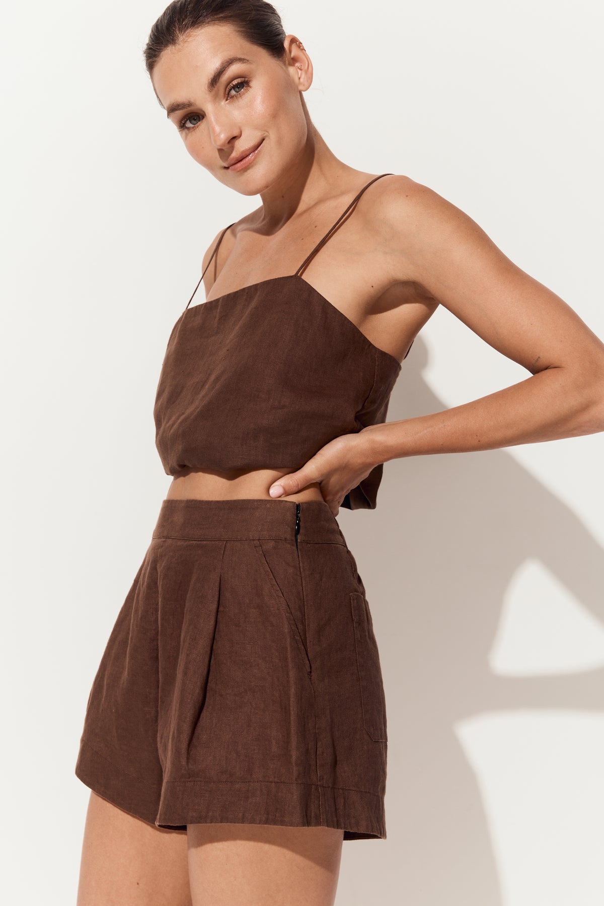 Gaia Linen Shorts in Chocolate - three quarter view, touching waistline with matching chocolate linen Rosa camisole - Lilly Pilly Collection