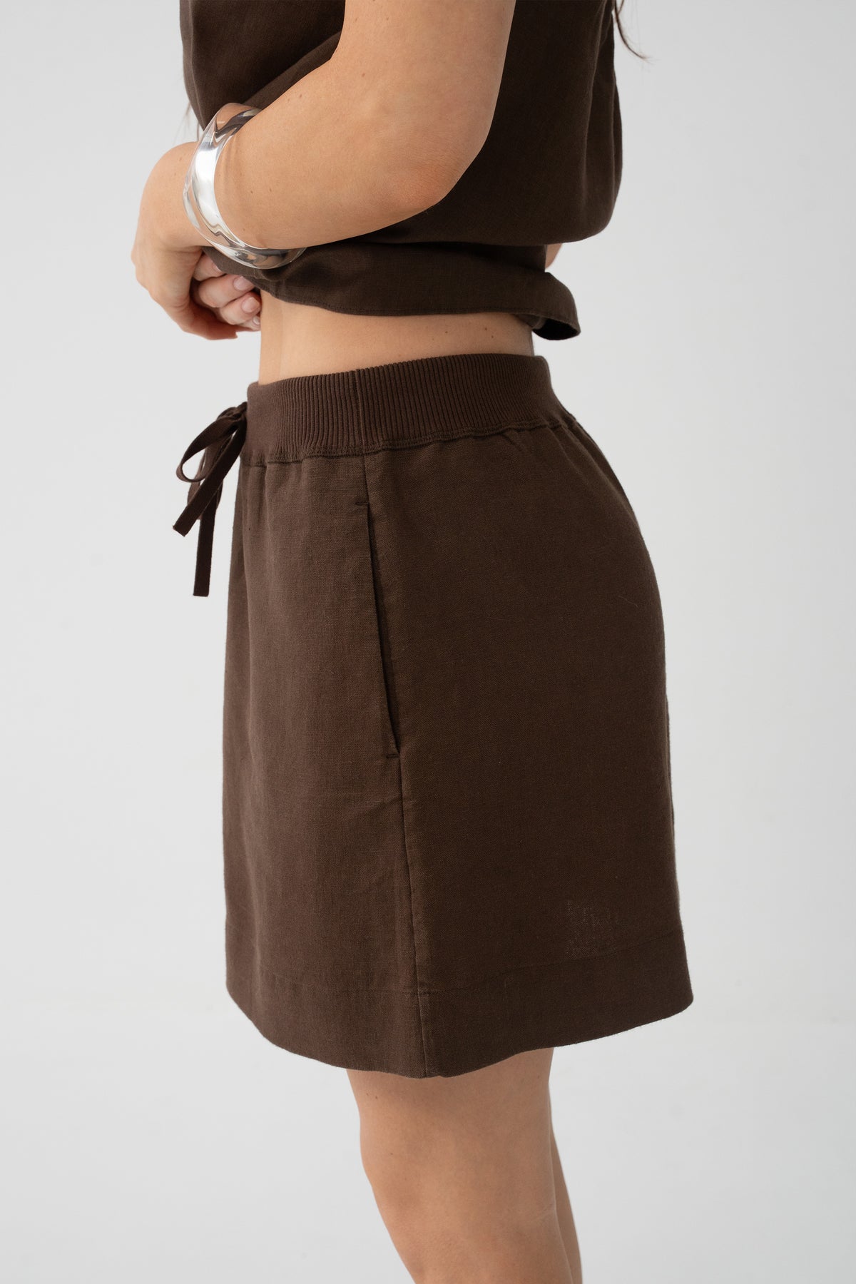 Anelle Linen Mini Skirt in Chocolate - side view, drawstring waistband, arms crossed - Lilly Pilly Collection