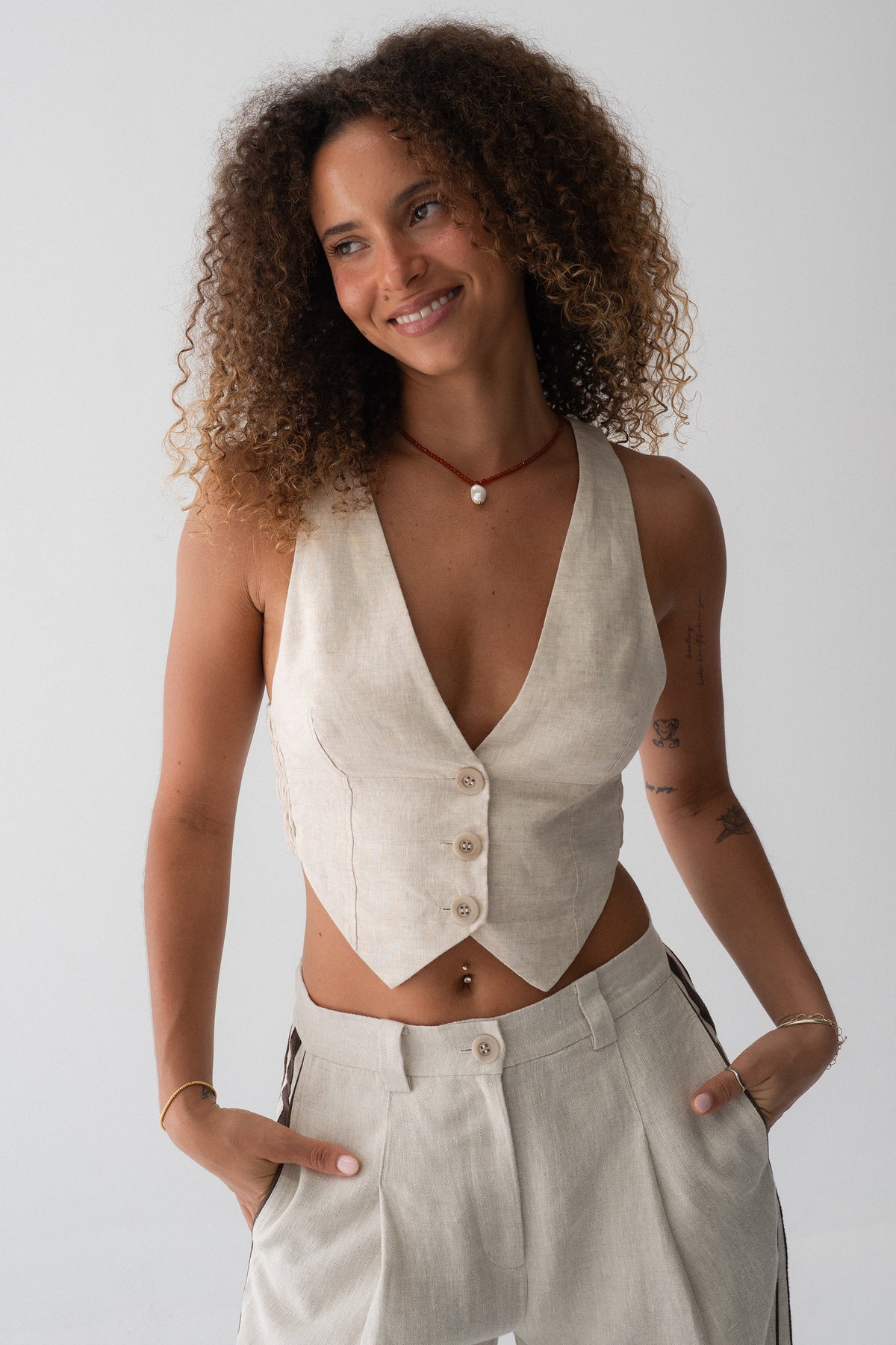 Ari Linen Vest in Oatmeal - front view, smiling, V-neckline and button-front with pointed hem, styled with wide-leg linen pants - Lilly Pilly Collection