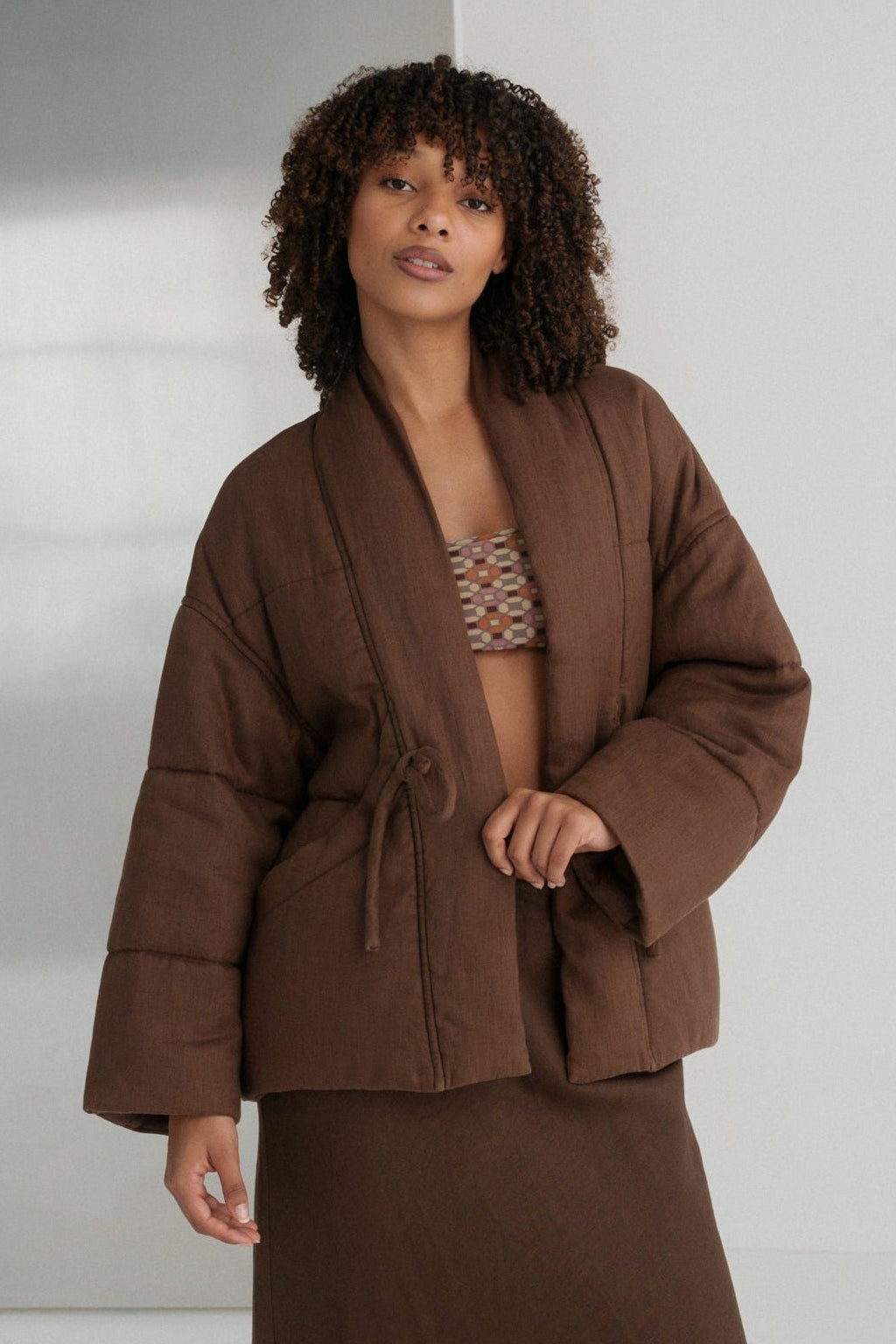 LILLY PILLY woman wearing a chocolate brown Aster linen jacket and Riley skirt standing against a white wall with a metallic object.