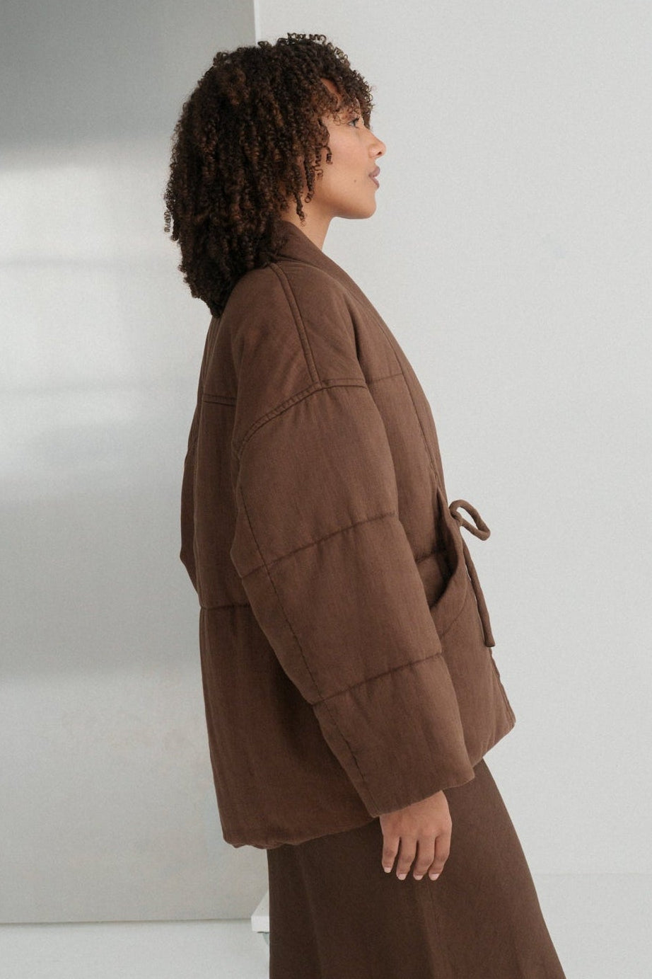 LILLY PILLY woman wearing a chocolate brown Aster linen jacket standing against a white wall.