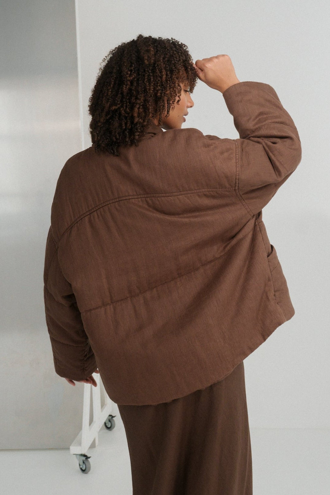 LILLY PILLY woman wearing a chocolate brown Aster linen jacket standing in a minimalistic room with white walls and a gray floor.