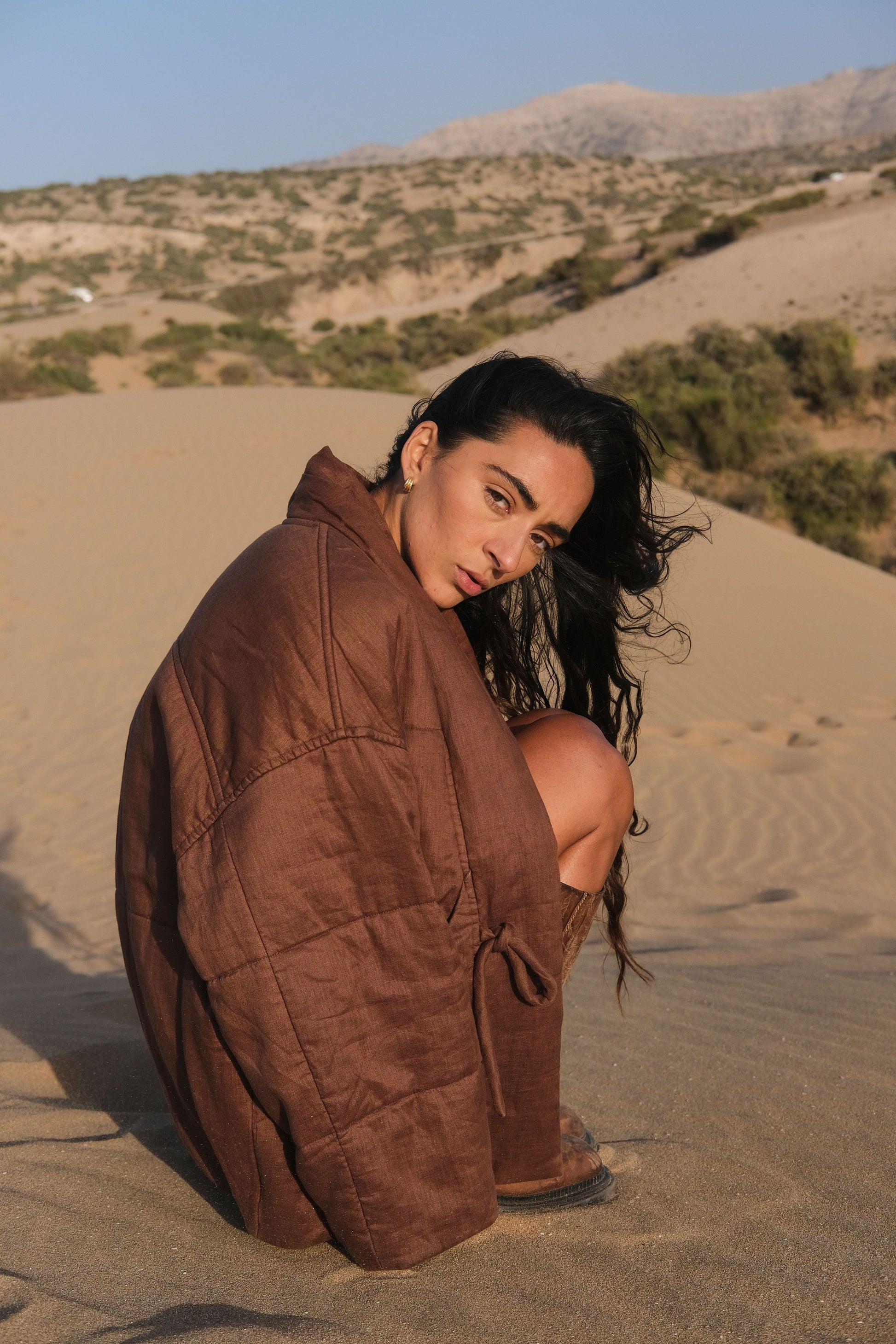 LILLY PILLY woman wearing a chocolate brown Aster linen jacket sitting on sand with mountains in the background