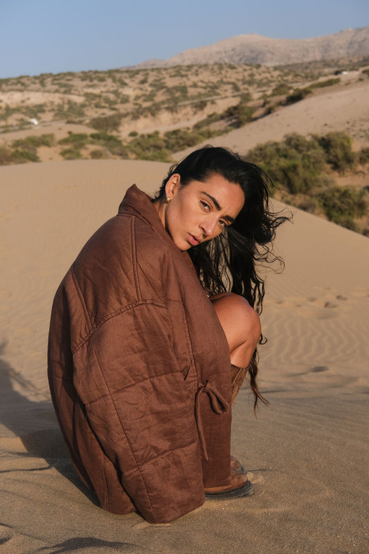 LILLY PILLY woman wearing a chocolate brown Aster linen jacket sitting on sand with mountains in the background