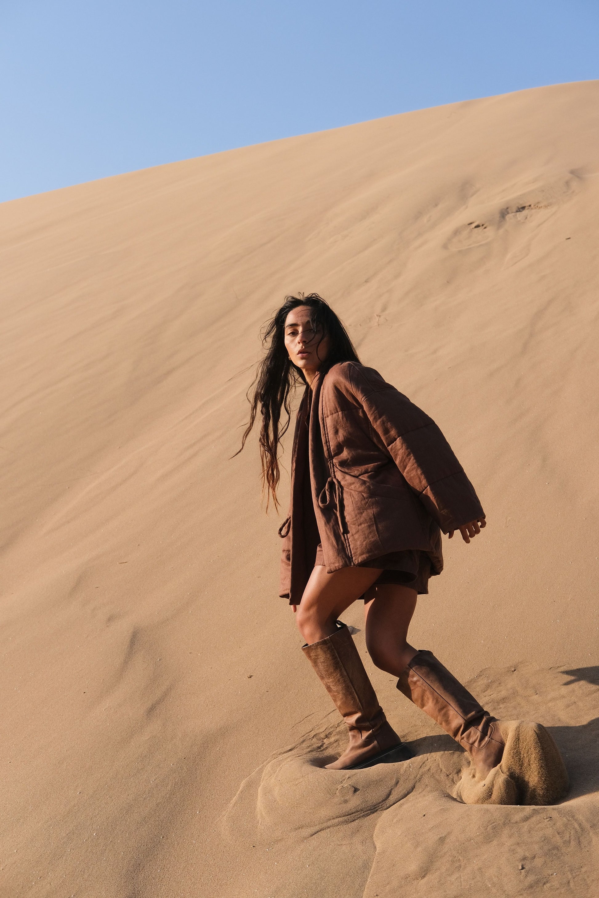LILLY PILLY woman wearing a chocolate brown Aster linen jacket and boots standing on a sand dune with a clear blue sky.