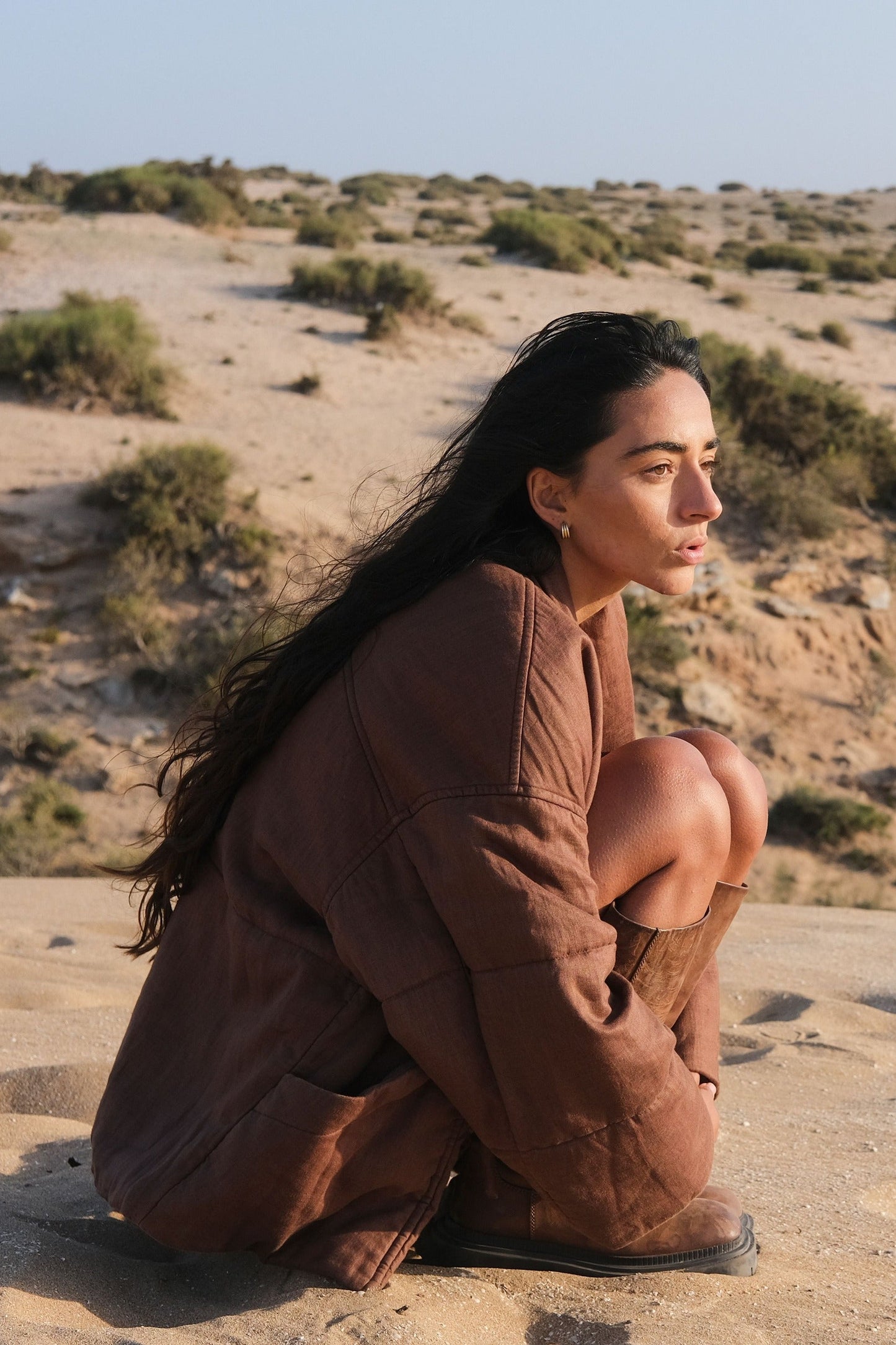 LILLY PILLY woman wearing a chocolate brown Aster linen jacket sitting on a sand dune in a desert landscape