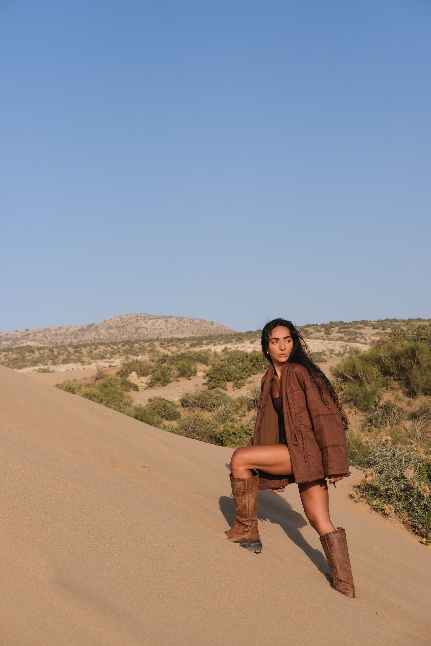 LILLY PILLY woman wearing a chocolate brown Aster linen jacket and boots standing on a sand dune with a clear blue sky.