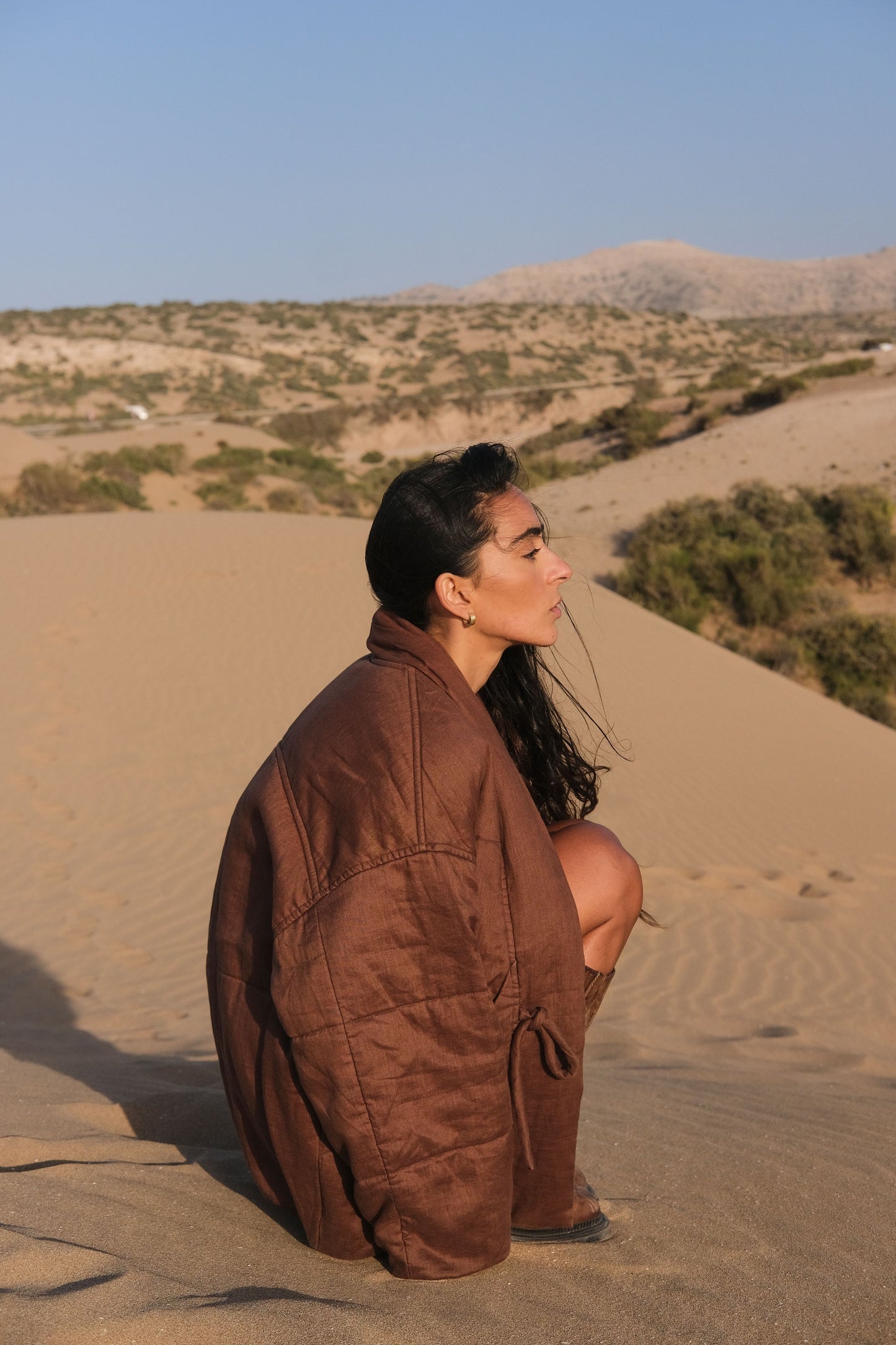 LILLY PILLY woman wearing a chocolate brown Aster linen jacket sitting on sand dunes with a desert landscape in the background