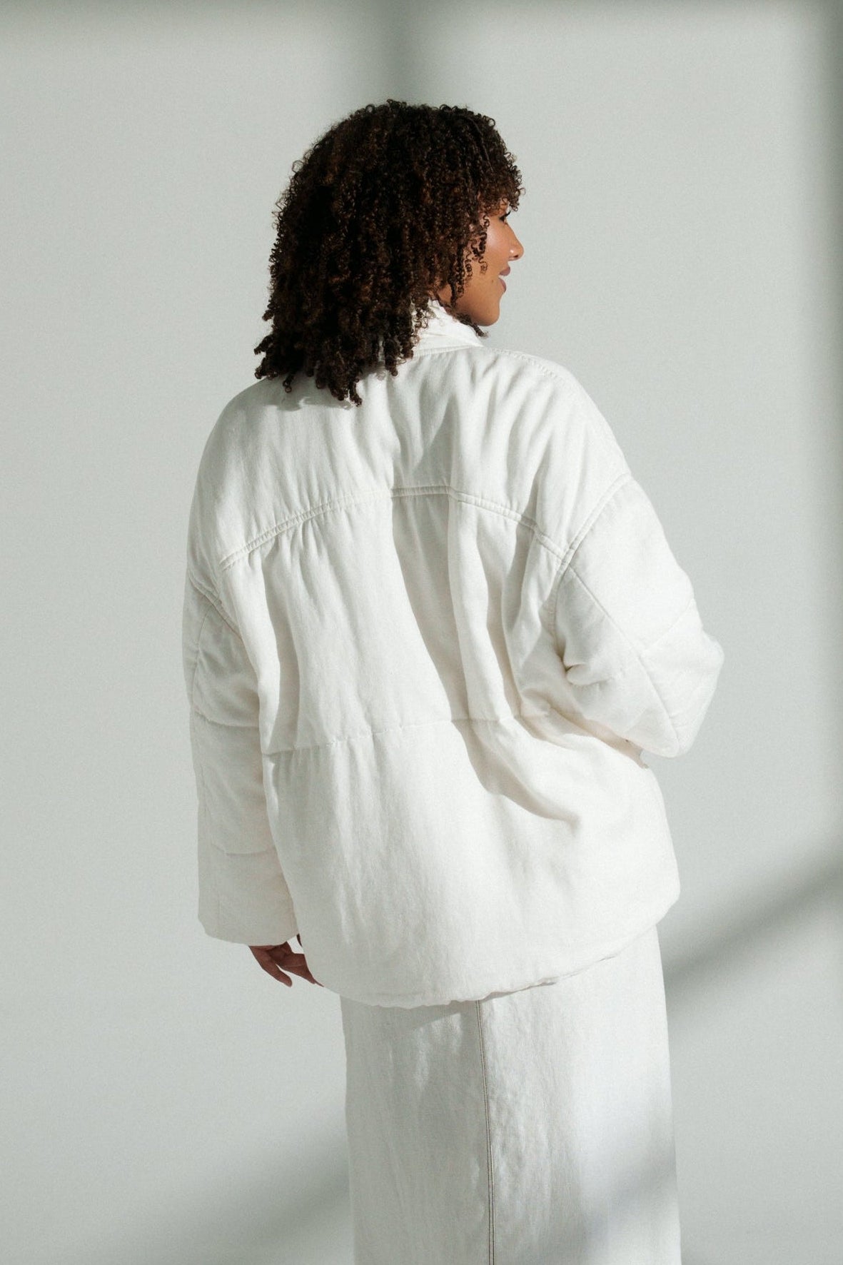 LILLY PILLY woman wearing Aster linen coat in ivory standing in a softly lit room with shadows on the wall.