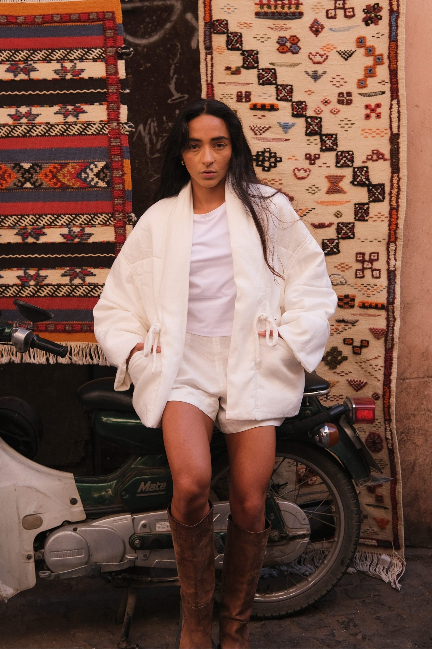 LILLY PILLY woman wearing Aster linen coat in ivory standing next to a scooter with patterned rugs in the background