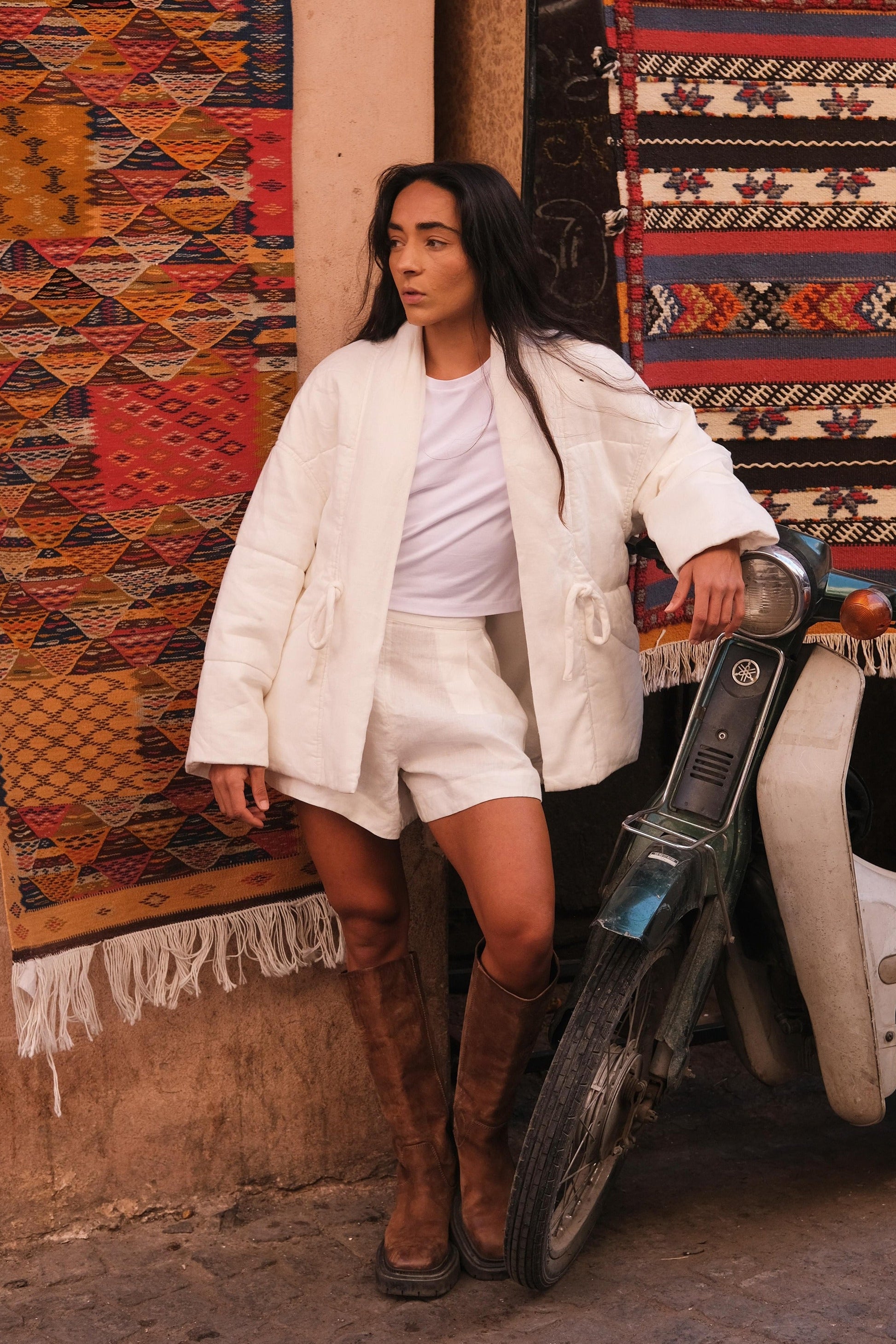 LILLY PILLY woman wearing Aster linen coat in ivory standing next to a scooter with patterned rugs in the background