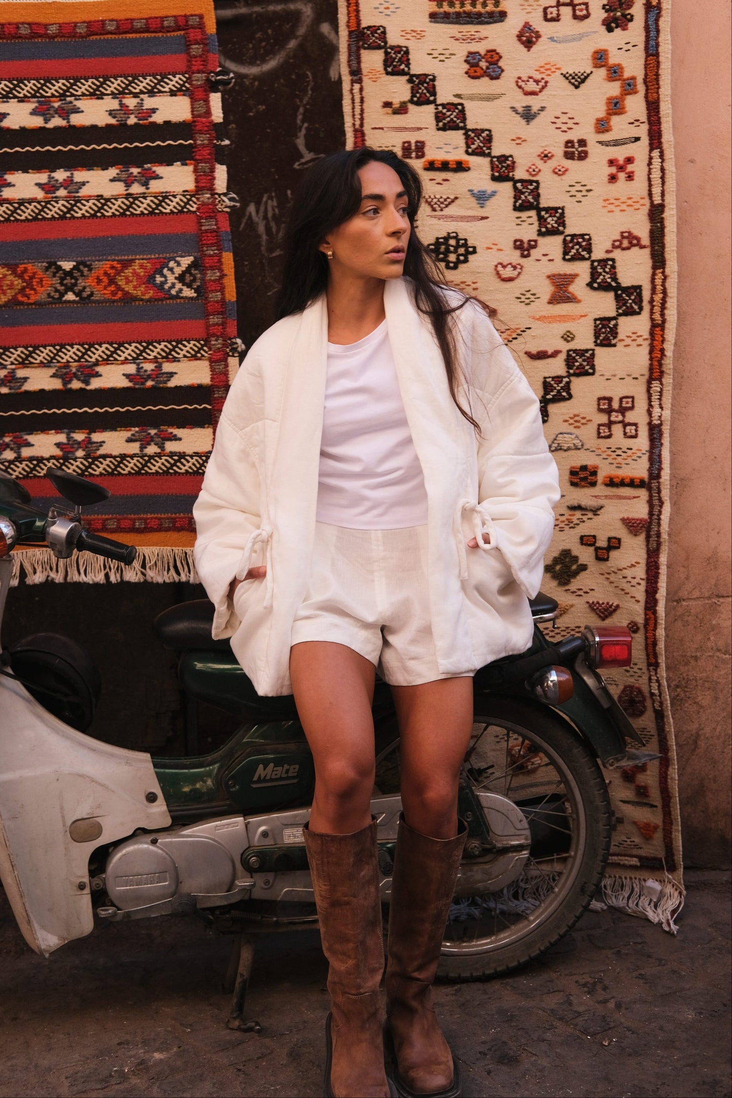 LILLY PILLY woman wearing Aster linen coat in ivory with brown boots standing next to a scooter with patterned rugs in the background