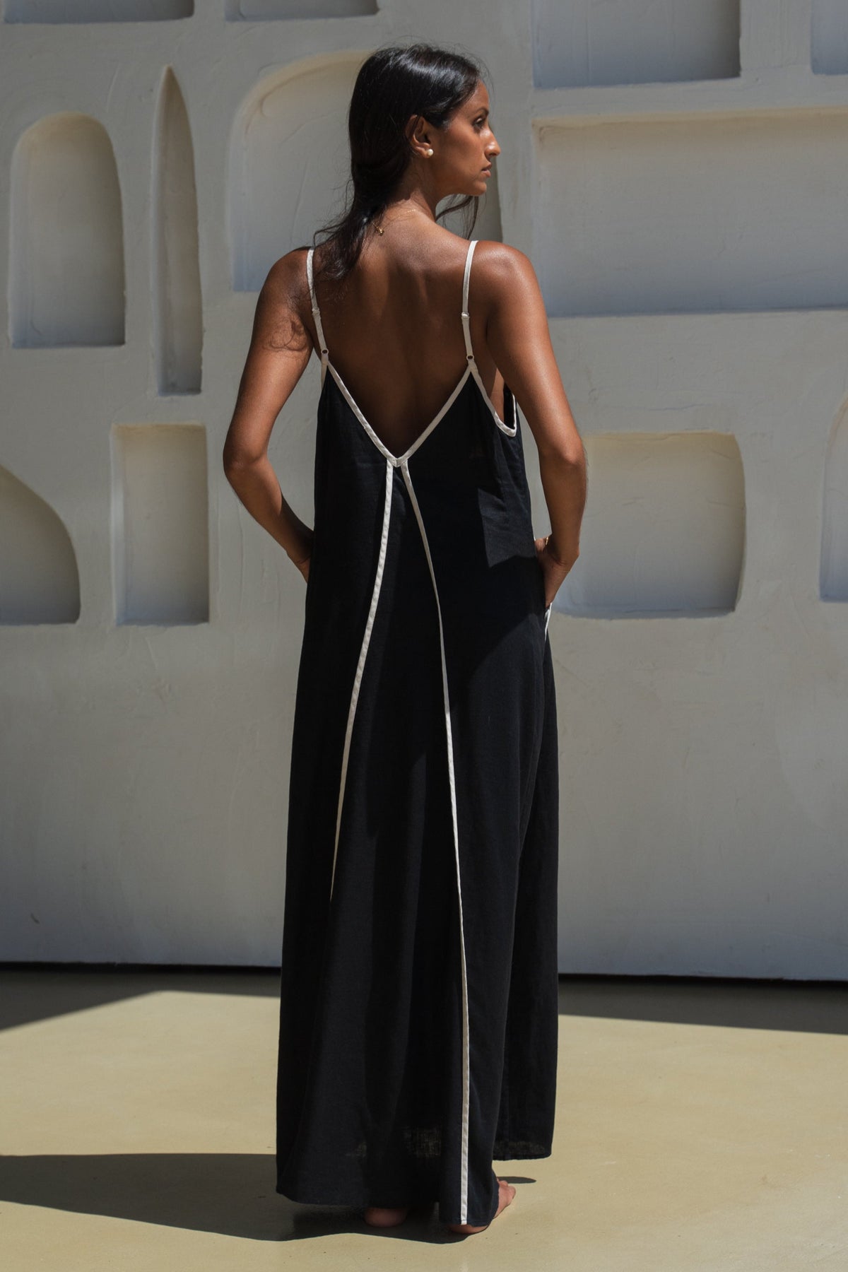 Back view of LILLY PILLY woman in a Azalea black linen dress with oatmeal trim standing in front of a white wall with geometric shapes