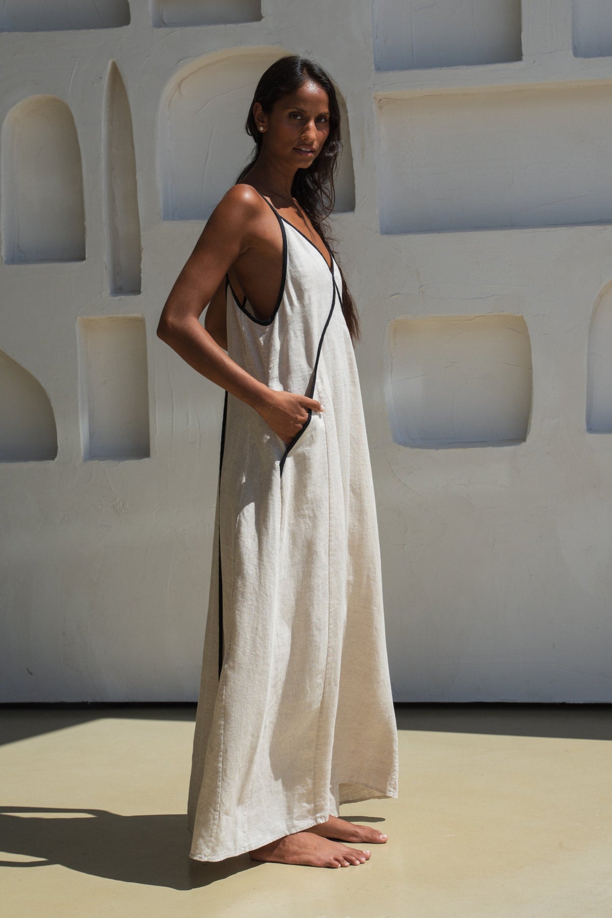 lilly pilly woman wearing an oatmeal Azalea dress with black straps against a light-colored wall showing the side view. 