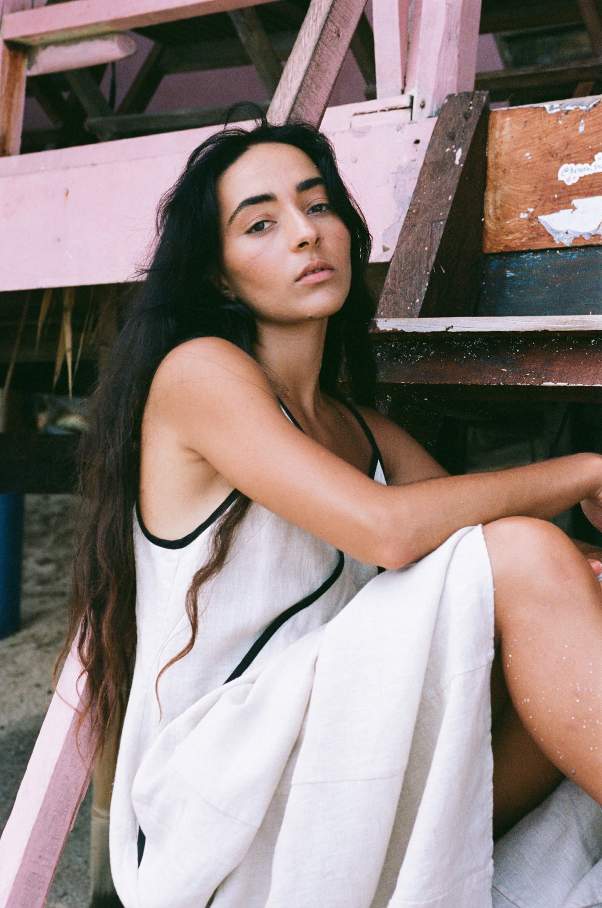 Lilly Pilly woman wearing an oatmeal Azalea dress with black straps and trim sitting on a wooden staircase wearing a white dress with black trim.