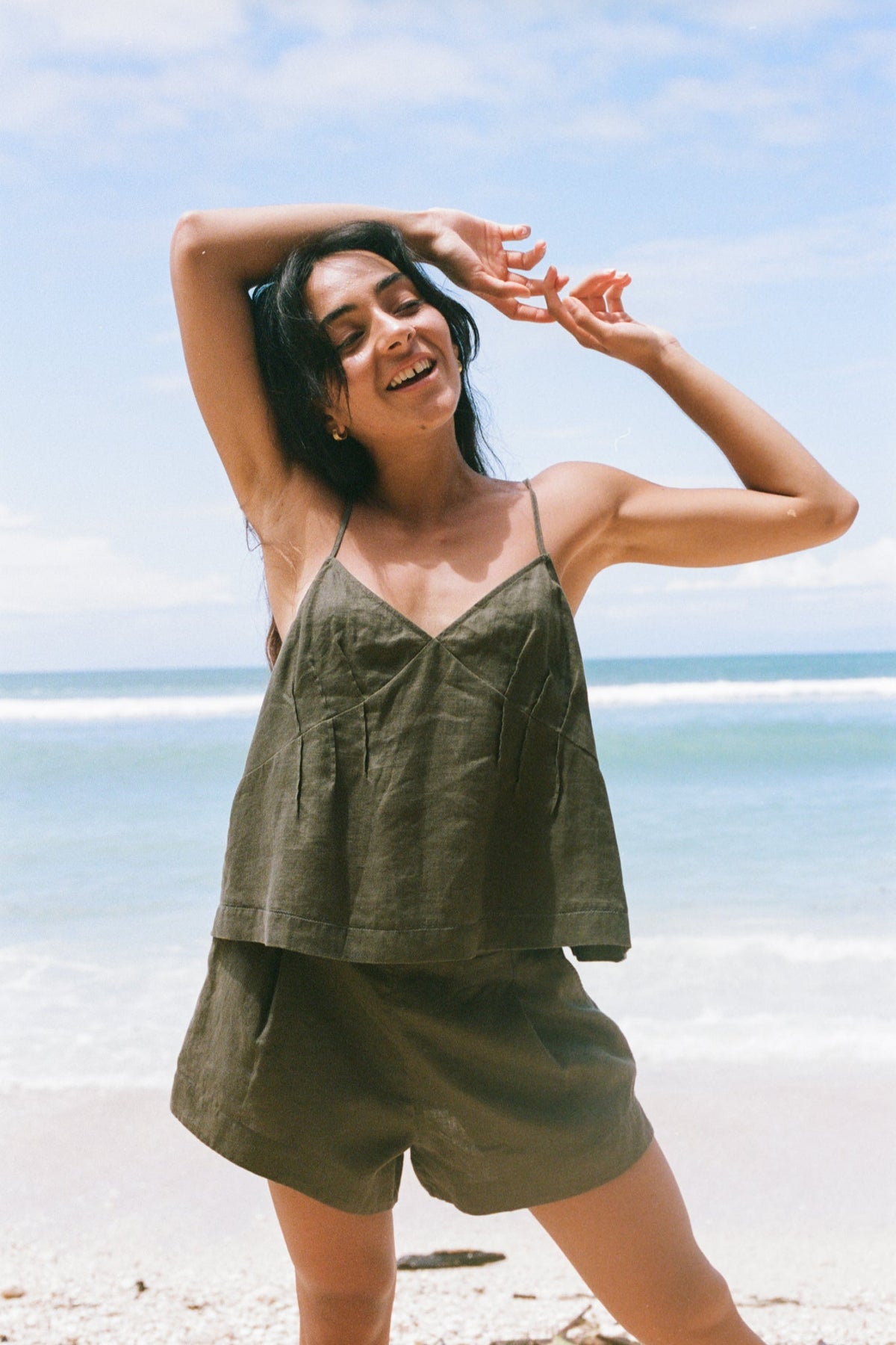 LILLY PILLY woman wearing a khaki Bea linen cami top and Gaia shorts outfit on a beach with clear blue sky holding her hands above her head