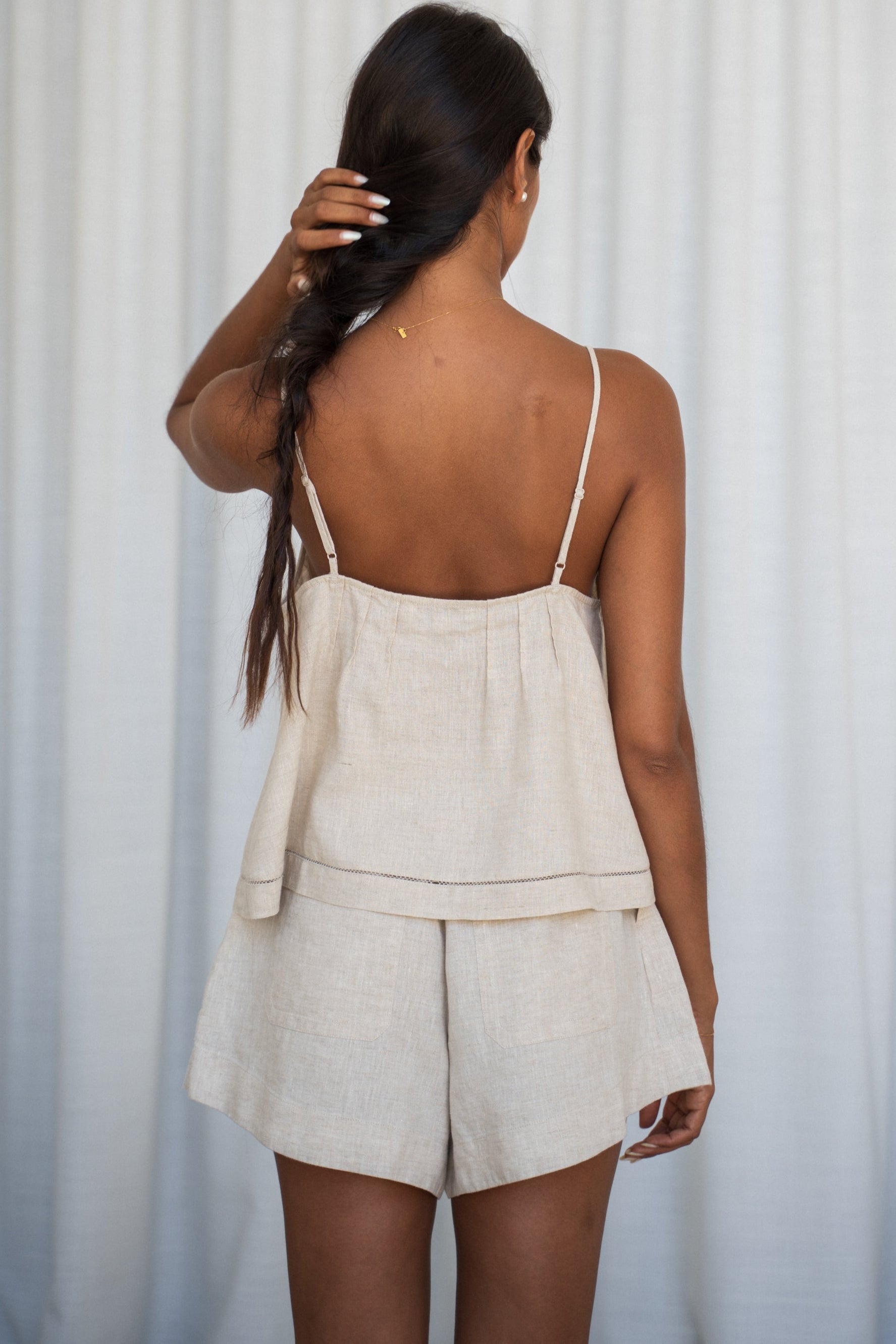 LILLY PILLY woman wearing a oatmeal Bea linen cami top and Gaia shorts outfit showing back view against a white curtain background
