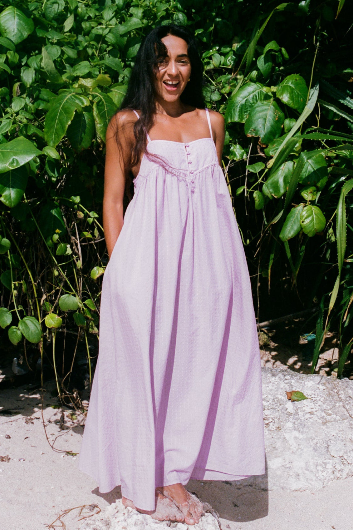 Woman in a light purple dress standing in a natural setting with greenery.