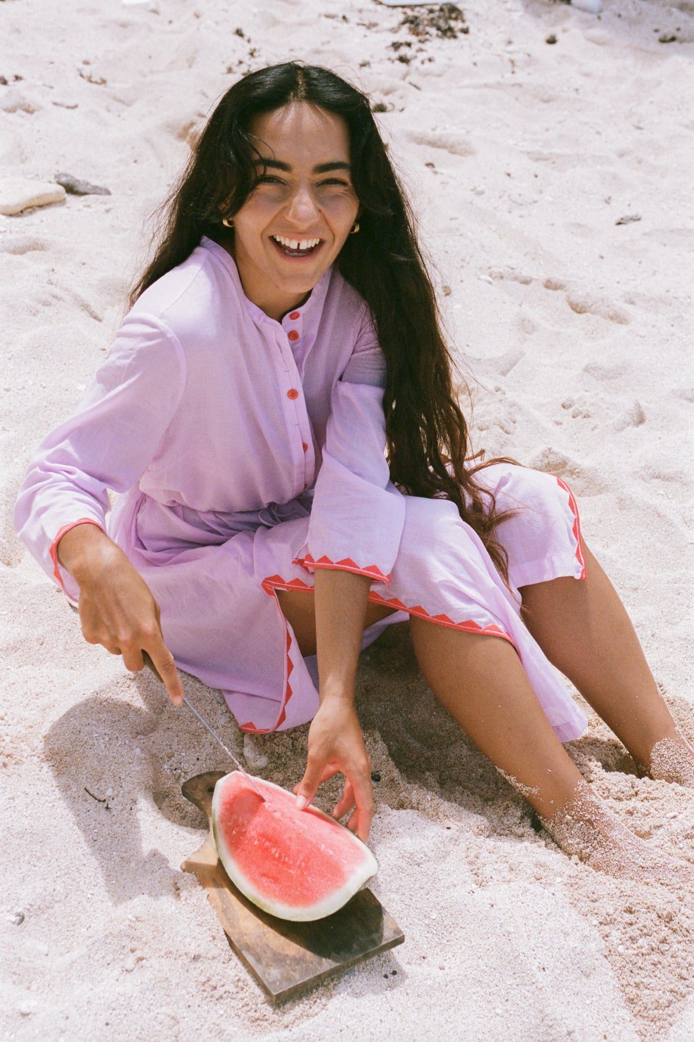 LILLY PILLY woman in an orchid Callie cotton dress sitting in the sand with a watermelon slice