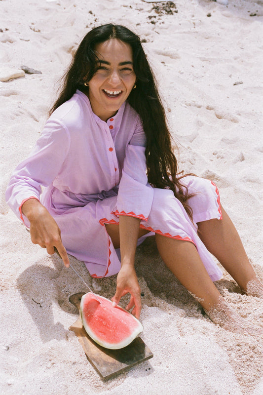 LILLY PILLY woman in an orchid Callie cotton dress sitting in the sand with a watermelon slice