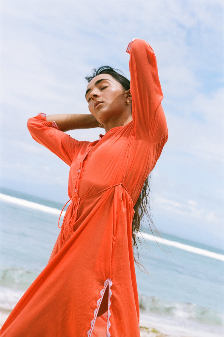Callie Cotton Voile Midi Dress in Spritz Orange - arms raised overhead, button placket and tie waist detail, sky and ocean backdrop - Lilly Pilly Collection