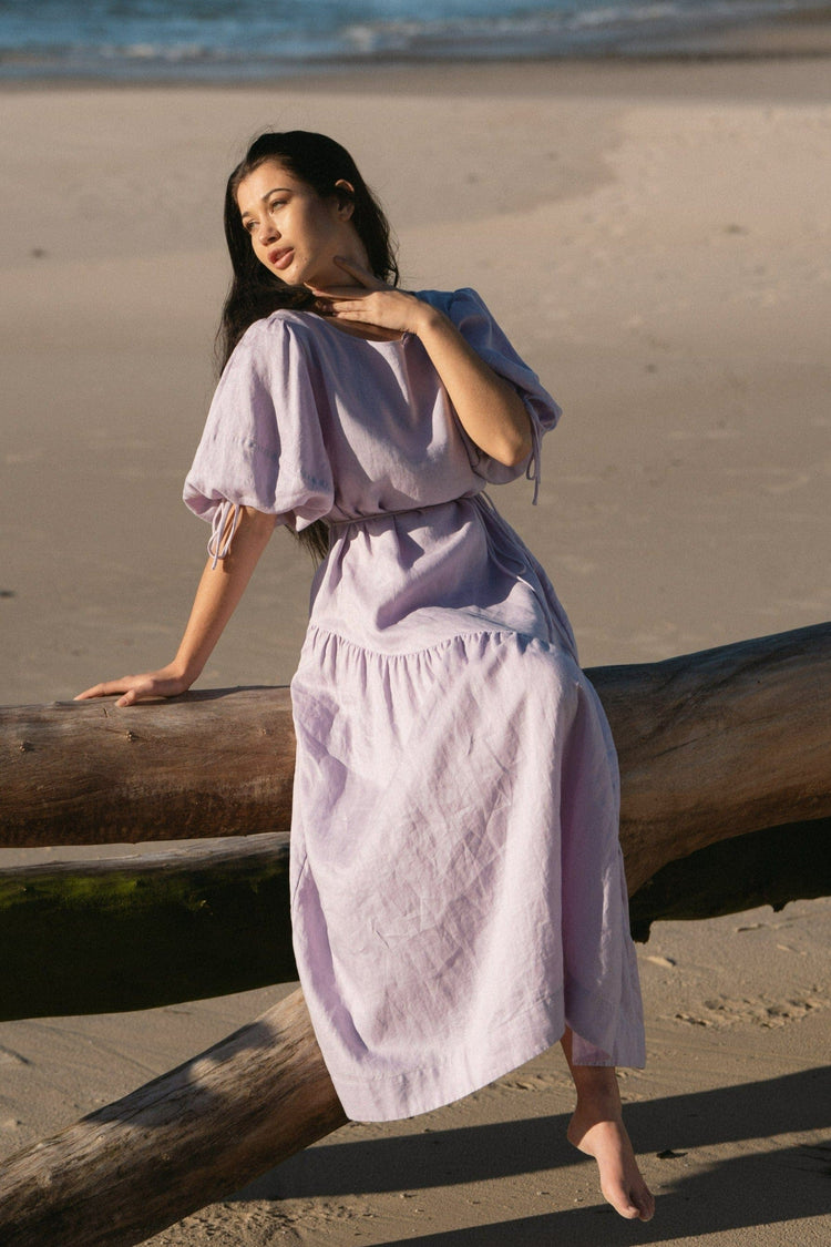 Charlotte Linen Maxi Dress in Lilac - sitting on driftwood, round neckline and tie-cuff puff sleeves, tiered skirt, beach at sunset - Lilly Pilly Collection