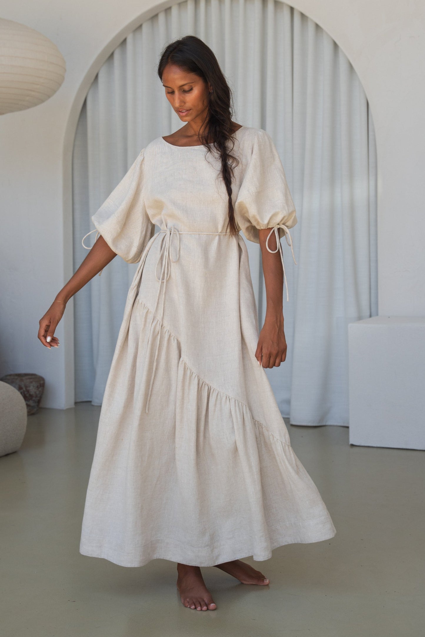 LILLY PILLY woman in an oatmeal Charlotte linen dress twirling around in a minimalistic room with a lamp and shelves.