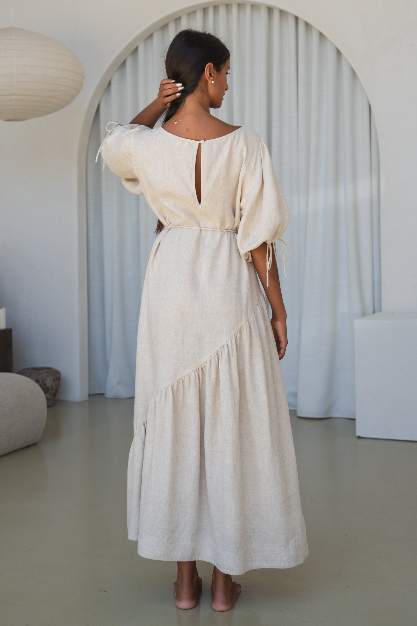 LILLY PILLY woman in an oatmeal Charlotte linen dress back view while standing in a minimalistic room with a lamp and shelves.