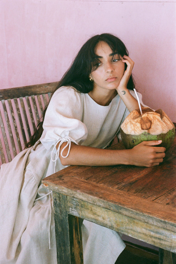 Charlotte Linen Maxi Dress in Oatmeal - side view sitting on a chair in a room with pink walls and a wooden table - Lilly Pilly Collection