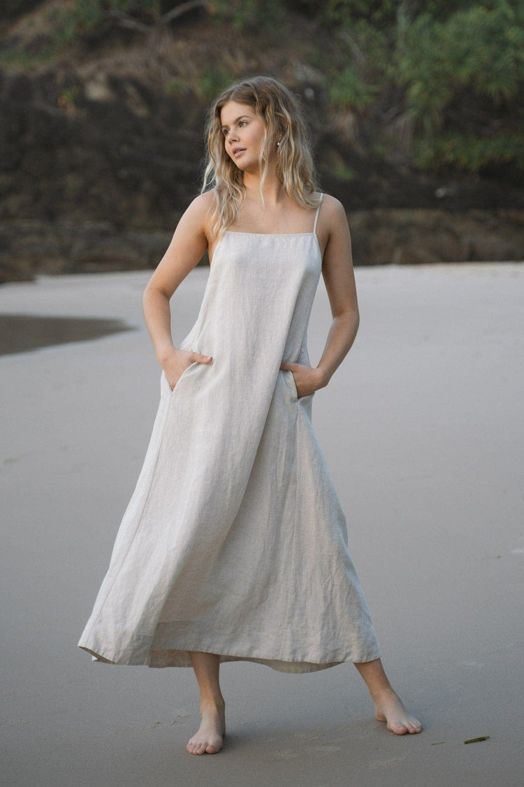 Coco Linen Midi Dress in Oatmeal - full length front view, hands in pockets, square neckline and spaghetti straps, standing on beach with coastal greenery backdrop - Lilly Pilly Collection