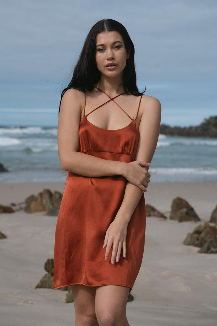 Ella Silk Mini Slip Dress in Rust Red - full length front view, hands crossed, standing on beach with rocks backdrop - Lilly Pilly Collection