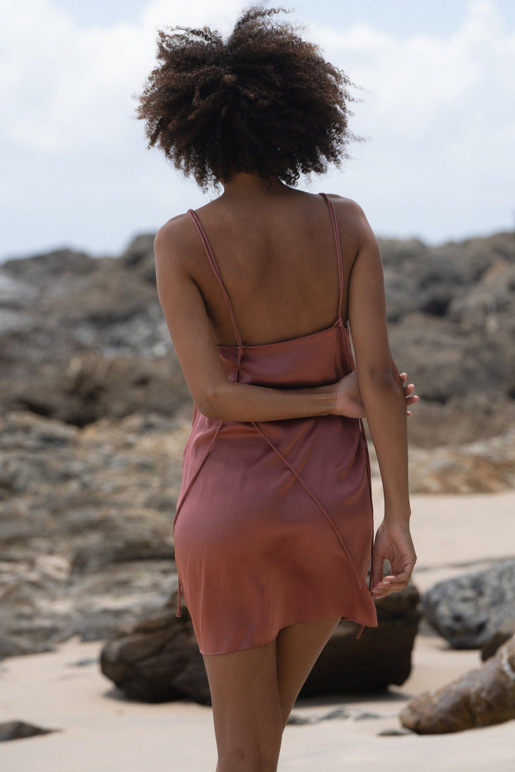 Ella Silk Mini Slip Dress in Dusty Pink - back view, criss-cross tie-strap detail, standing on beach with rocks backdrop - Lilly Pilly Collection