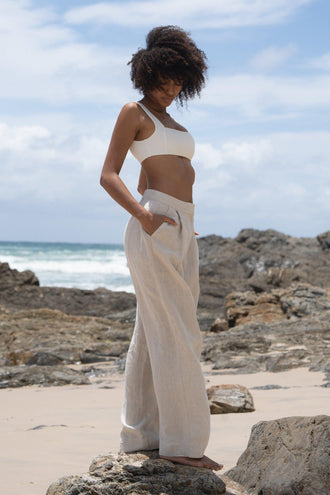 Oli Linen Pants in Oatmeal - side view showing front pleat and wide-leg linen silhouette, woman wearing ivory square-neck crop bra top, rocky beach backdrop - Lilly Pilly Collection