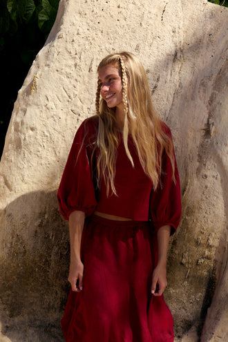 Demi Linen Top in Cherry Red - woman smiling against sandstone wall, round neckline and puff sleeves, styled with matching Riley Linen Skirt - Lilly Pilly Collection