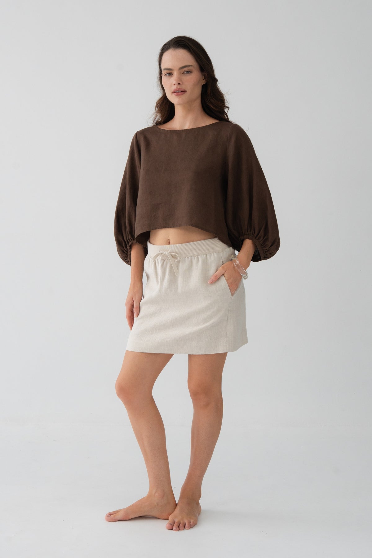 Demi Linen Top in Chocolate - three-quarter view, hand in pocket, puff sleeves and cropped hem, styled with oatmeal linen mini skirt - Lilly Pilly Collection