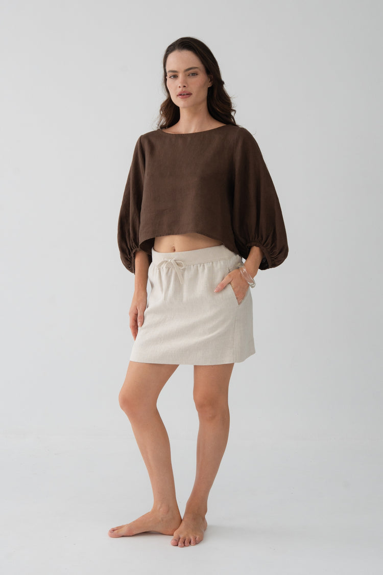 Demi Linen Top in Chocolate - three-quarter view, hand in pocket, puff sleeves and cropped hem, styled with oatmeal linen mini skirt - Lilly Pilly Collection