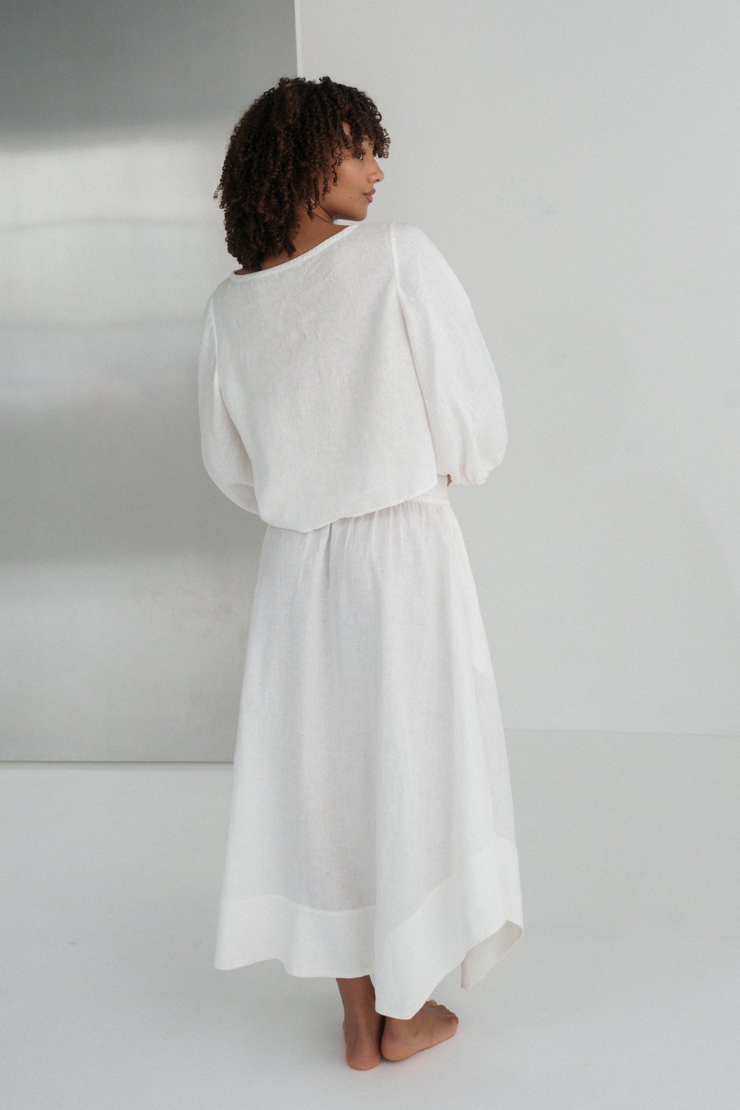 LILLY PILLY woman wearing an ivory Demi linen top and Hannah skirt standing in a minimalistic room with a large mirror.