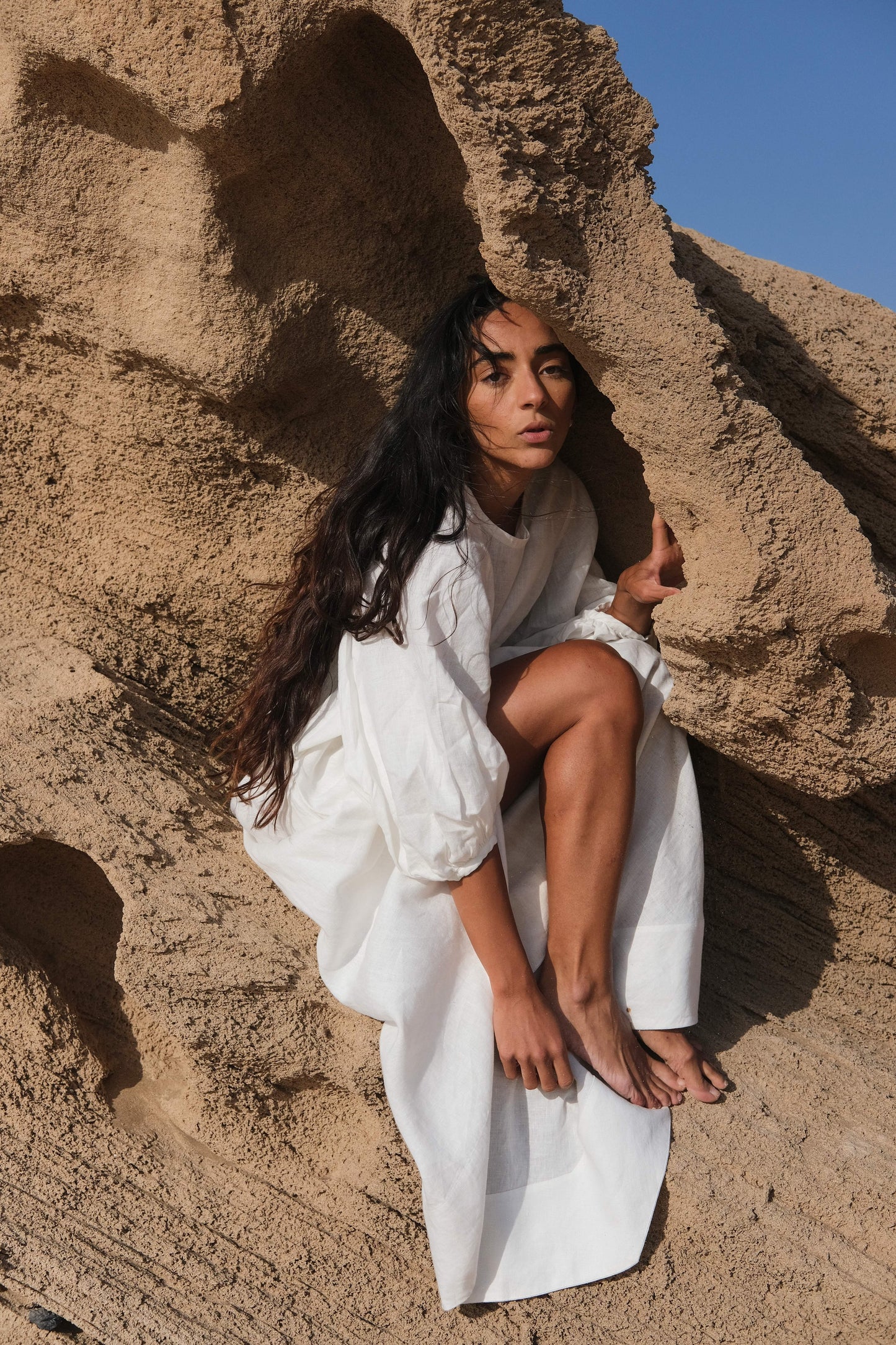 LILLY PILLY woman wearing an ivory Demi linen top and Hannah skirt sitting against a sandstone wall with a clear blue sky.