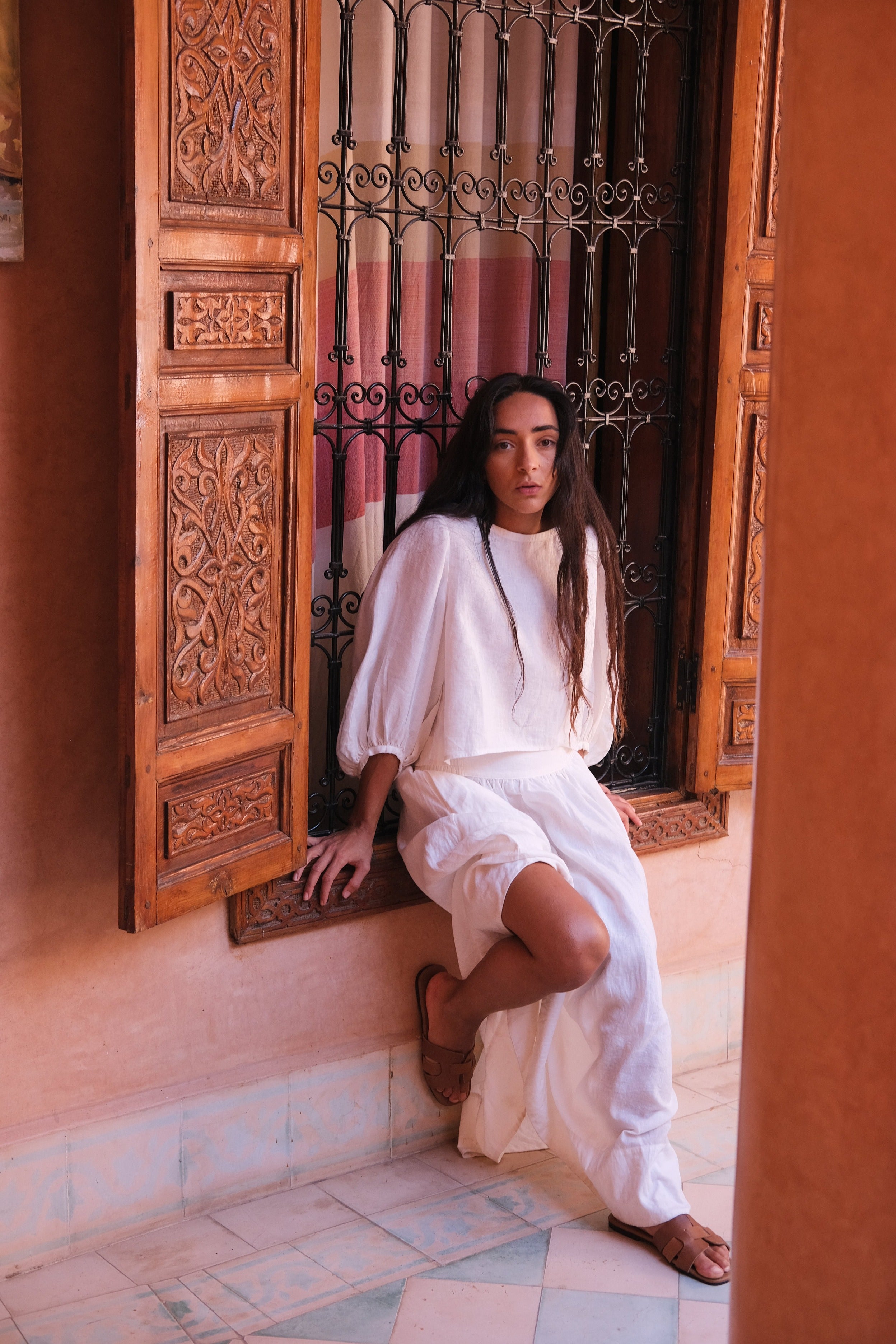 LILLY PILLY woman wearing an ivory Demi linen top and Hannah skirt sitting in a room with ornate wooden doors and a tiled floor.