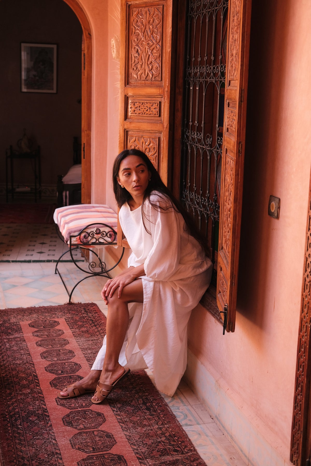 LILLY PILLY woman wearing an ivory Demi linen top and Hannah skirt sitting on a chair in a room with pink walls and decorative elements.