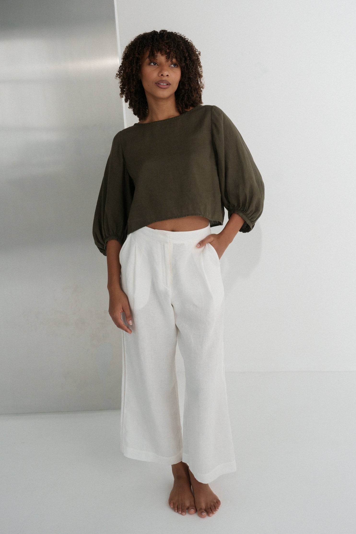 LILLY PILLY woman wearing a khaki green Demi linen top and ivory Ivy linen pants standing against a plain wall.